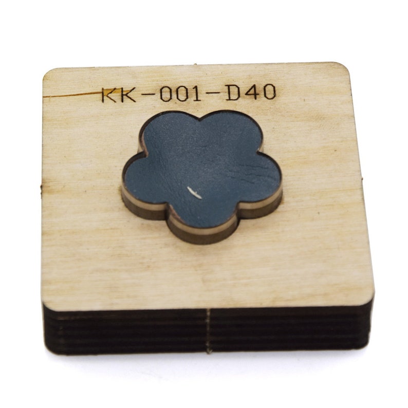 May include: A wooden die-cut tool with a flower-shaped cutout. The flower is dark blue and sits on a square wooden base. The text "KK-001-D40" is printed on the wood. The tool is likely used for crafting or leatherwork.