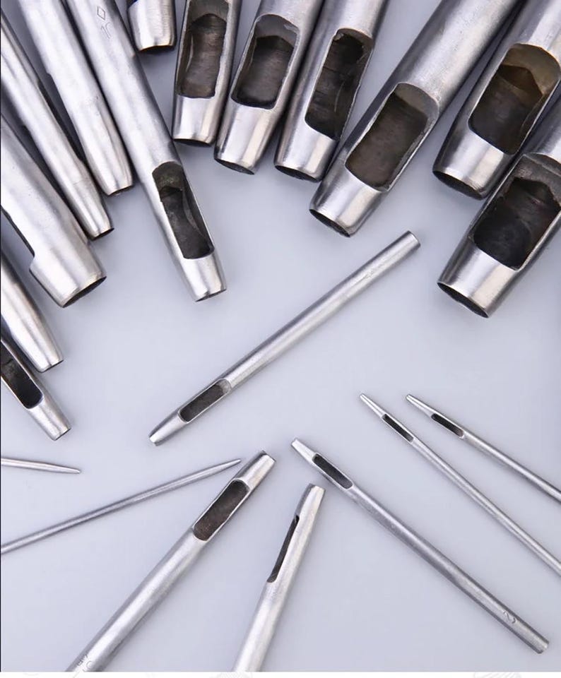 May include: A collection of silver metal leather working tools, including various sizes of hole punches and awls, arranged on a light gray surface. The tools are designed for creating precise holes in leather and similar materials.