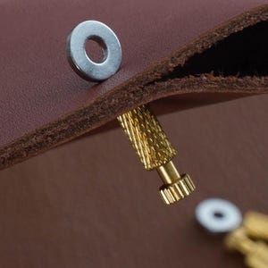 May include: Close-up of a brown leather item with a silver metal rivet and a gold-coloured screw. The leather has a textured appearance. The screw is partially inserted into the leather.