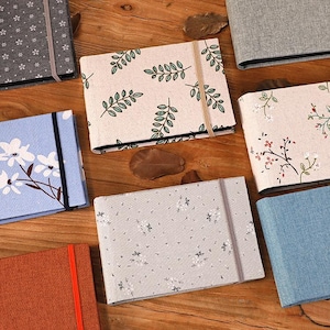 May include: A collection of rectangular journals with floral and botanical patterns. The journals feature various colors, including blue, gray, and orange, and are secured with elastic bands. The covers display leaf and flower designs.