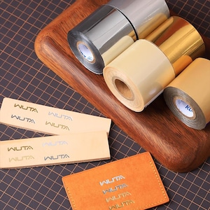 May include: A selection of metallic foil rolls in silver, gold, and beige, with small leather pieces and a card holder, all branded with the "WUTA" logo. The items are displayed on a wooden board and a dark grid surface.