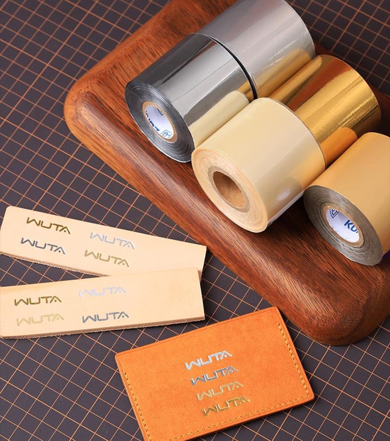 May include: A selection of metallic foil rolls in silver, gold, and beige, with small leather pieces and a card holder, all branded with the "WUTA" logo. The items are displayed on a wooden board and a dark grid surface.