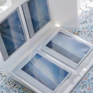 May include: A white photo album with several photos of a seascape. The album is open, displaying multiple images in clear plastic sleeves. The photos feature a blue sky and ocean. The album is on a floral patterned surface.