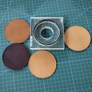 May include: A leathercrafting setup featuring a clear acrylic circle cutter with a black ring, surrounded by five circular leather pieces in shades of brown and purple. The items are arranged on a green grid surface.