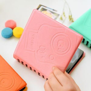 May include: A pink spiral-bound notebook with an embossed camera design and the word "instax". The notebook is open, revealing black pages. Colorful macarons are in the background. Other notebooks in orange and green are also visible.