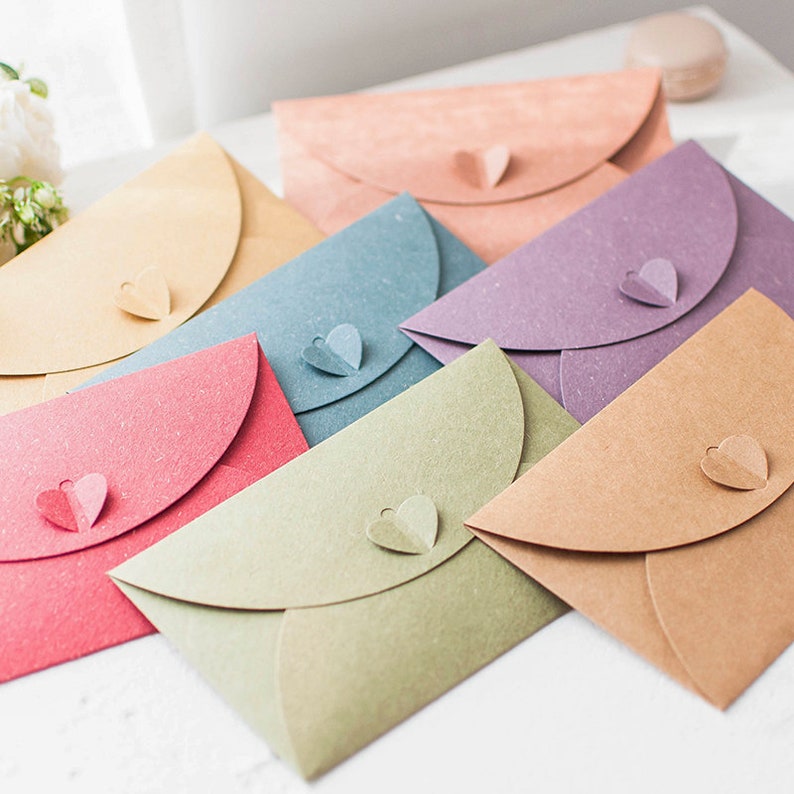 May include: A collection of colorful paper envelopes in shades of pink, blue, green, purple, and tan. Each envelope features a heart-shaped closure. The envelopes are arranged in a slightly overlapping pattern, suggesting a stationery or craft theme.