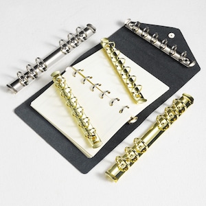 May include: A selection of planner binder rings in silver and gold. The rings are positioned around a planner with cream-coloured pages and a dark grey cover. The rings are designed to secure the pages.