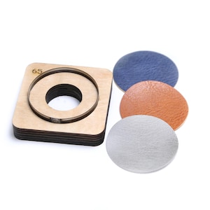 Puede incluir: Una herramienta de corte de madera con un centro circular, etiquetada como "65", junto a tres círculos de cuero en azul, naranja y plateado. La herramienta de corte es cuadrada con un recorte circular. Los círculos de cuero tienen un aspecto texturizado.