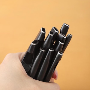 May include: A collection of black leather hole punches with silver-colored rims, held in a hand against a warm, orange background. The tools have various shaped cutting ends, designed for leathercraft and similar projects.