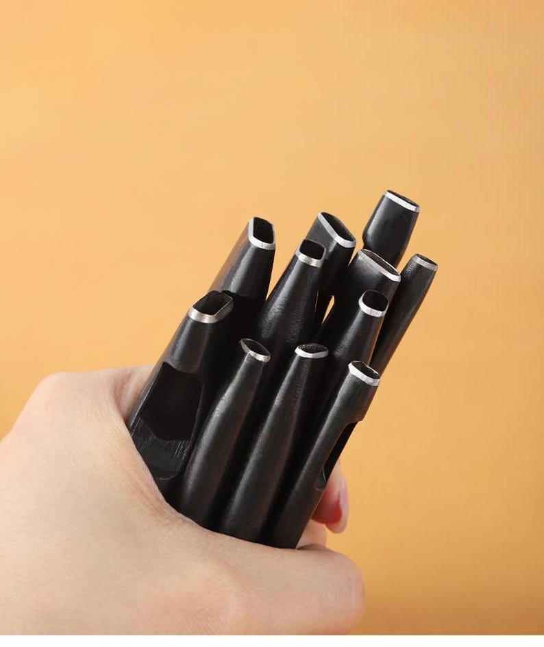 May include: A collection of black leather hole punches with silver-colored rims, held in a hand against a warm, orange background. The tools have various shaped cutting ends, designed for leathercraft and similar projects.