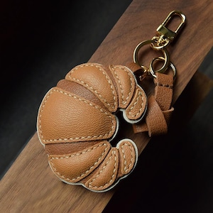 May include: A brown leather croissant keychain with gold-colored hardware. The keychain features a detailed stitched design and a small leather strap. The croissant is a light brown color with darker brown accents.