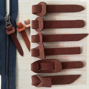 May include: A selection of brown suede zipper pulls and tabs in various shapes. A dark blue zipper is visible on the left. The tabs are in shades of brown and orange, suitable for clothing or accessories.