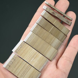May include: A collection of small, rectangular metal blades with sharp edges, arranged in a hand. The blades have a light brown color with a silver edge. Some blades have text printed on them. The background is a solid dark gray.