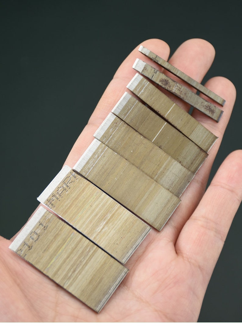May include: A collection of small, rectangular metal blades with sharp edges, arranged in a hand. The blades have a light brown color with a silver edge. Some blades have text printed on them. The background is a solid dark gray.
