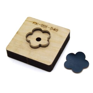 May include: A wooden flower-shaped die cut tool with the text "KK-001-D40" engraved on the surface. A dark blue flower-shaped cutout is next to the die cut. The die cut is made of stacked wooden layers.