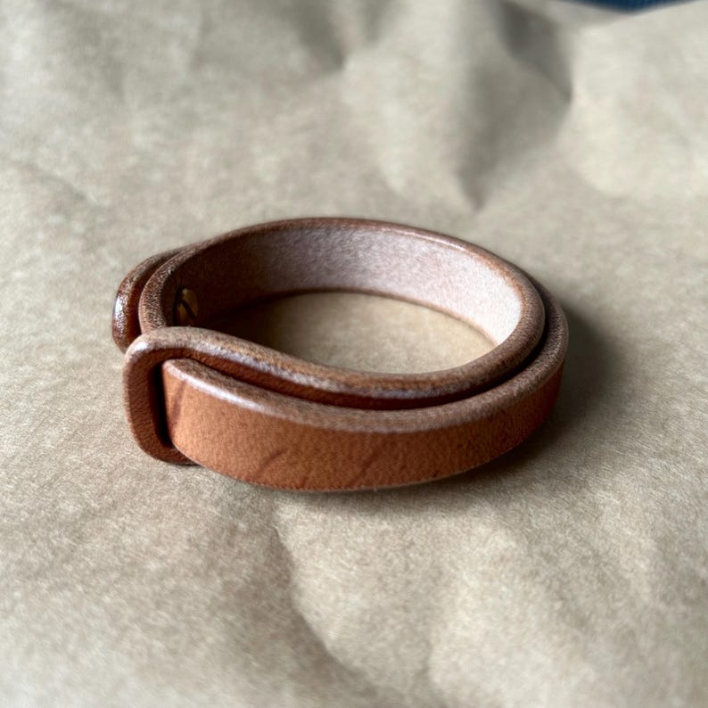 May include: A light brown leather bracelet with a buckle closure. The bracelet is made of two layers of leather and appears to be new. The background is a light brown, textured surface.