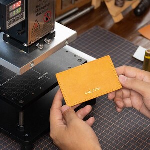 May include: A mustard leather cardholder with gold "WUTA" branding, held near a hot foil stamping machine. The machine displays digital readouts and warning labels. The cardholder is approximately 10 cm x 7 cm.
