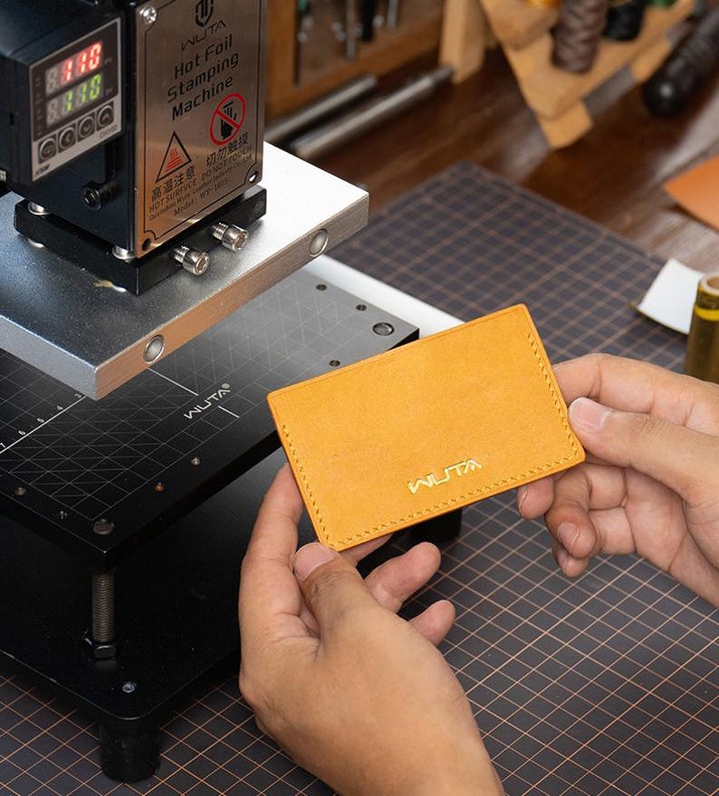 May include: A mustard leather cardholder with gold "WUTA" branding, held near a hot foil stamping machine. The machine displays digital readouts and warning labels. The cardholder is approximately 10 cm x 7 cm.