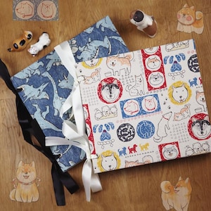 May include: Two square, fabric-covered journals with dog-themed patterns. One journal has a blue dinosaur print, the other features various dog breeds in colorful circles and squares. Both are tied with ribbons. Decorative dog figurines are scattered around.