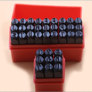 Puede incluir: Un juego de sellos de metal con letras y n&uacute;meros en una caja de pl&aacute;stico roja. Los sellos son negros y est&aacute;n dispuestos en filas. La caja est&aacute; abierta, revelando los sellos. El fondo es de color beige claro.