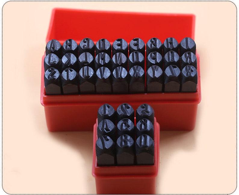 Puede incluir: Un juego de sellos de metal con letras y n&uacute;meros en una caja de pl&aacute;stico roja. Los sellos son negros y est&aacute;n dispuestos en filas. La caja est&aacute; abierta, revelando los sellos. El fondo es de color beige claro.