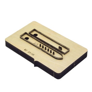 Pode incluir: Uma ferramenta de corte de madeira com base retangular e bordas em camadas. Duas matrizes de corte de metal são fixadas à superfície, uma com formato curvo e outra retangular. O número "2.5" é visível na matriz. O texto "BD 20-18" também é visível.