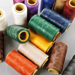 May include: A collection of spools of colorful thread in various shades, including white, purple, green, yellow, orange, beige, brown, and blue. The thread is tightly wound around cardboard cores, ideal for crafting and sewing projects.