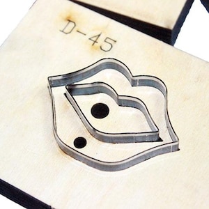 May include: A wooden die cut with a lip-shaped design. The die is made of light-colored wood and has a metal cutting edge. The text "D-45" is stamped on the wood. The die is likely used for crafting or leatherwork.