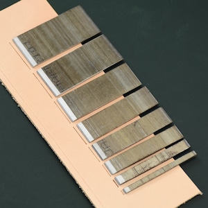 May include: A collection of metal leatherworking tools with sharp, silver blades, arranged on a piece of light brown leather. The tools vary in size, with etched markings on the blades. The background is a dark green surface.