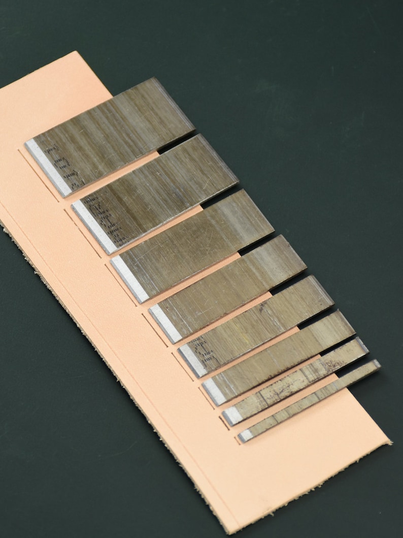 May include: A collection of metal leatherworking tools with sharp, silver blades, arranged on a piece of light brown leather. The tools vary in size, with etched markings on the blades. The background is a dark green surface.