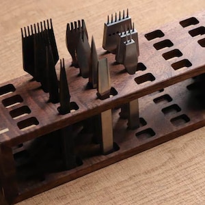 Puede incluir: Un organizador de herramientas de madera con varias herramientas de marroquinería. El organizador es rectangular con múltiples ranuras y compartimentos. Las herramientas incluyen cosedores de bordes, punzones y otros implementos de artesanía en cuero. La madera tiene un acabado marrón oscuro.