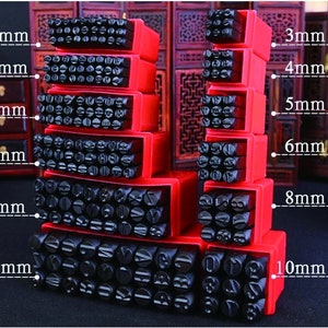 Puede incluir: Un conjunto de sellos de metal negro con letras y n&uacute;meros en cajas de almacenamiento rojas. Los tama&ntilde;os var&iacute;an de 3 mm a 10 mm. Los sellos est&aacute;n dispuestos en una exhibici&oacute;n apilada, mostrando varias fuentes y tama&ntilde;os.