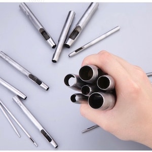 May include: A collection of silver-colored leather working tools, including hollow punches and needles, are displayed on a light gray surface. A hand holds a bundle of the hollow punches.