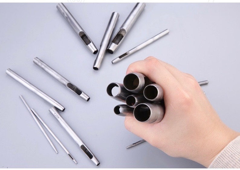 May include: A collection of silver-colored leather working tools, including hollow punches and needles, are displayed on a light gray surface. A hand holds a bundle of the hollow punches.