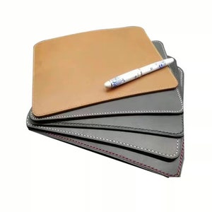 May include: A stack of rectangular leather desk pads in shades of brown and gray, with white stitching around the edges. A white pen with a blue floral design rests on top of the brown pad.
