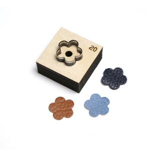 May include: A wooden flower-shaped die-cut tool with a metal cutting edge, alongside three leather flower cutouts in brown, blue, and navy. The number "20" is etched on the wooden block. Ideal for leathercraft and crafting projects.