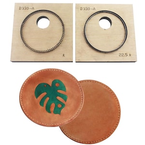 Può includere: Due strumenti artigianali in legno con disegni circolari e un foro al centro, etichettati "D330-A" e "22.5". Sotto, sono visibili due sottobicchieri in pelle marrone, uno con un disegno a foglia verde.