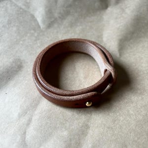 May include: A brown leather wrap bracelet with a gold-colored stud closure. The bracelet is wrapped twice around, showcasing the texture and color of the leather. The bracelet is approximately 7 inches in length.