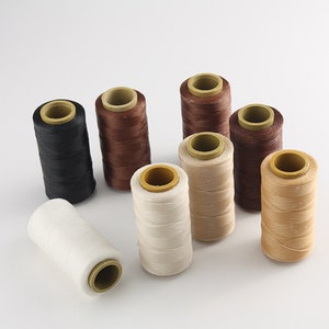 May include: A collection of spools of thread in various colors, including black, brown, beige, and white. The thread is wound around cardboard cores with yellow centers. The spools are arranged on a white background.