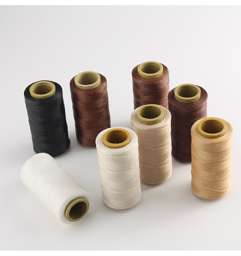 May include: A collection of spools of thread in various colors, including black, brown, beige, and white. The thread is wound around cardboard cores with yellow centers. The spools are arranged on a white background.