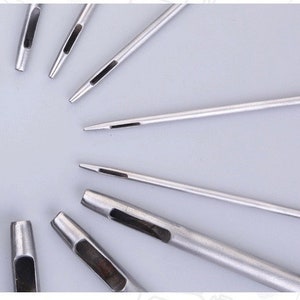 May include: A collection of silver metal leather working tools, including various sizes of hole punches and needles, arranged on a light gray surface. The tools are designed for creating precise holes and stitching in leather.