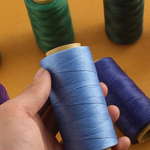 May include: A hand holds a spool of light blue thread, with other spools of green and purple thread in the background. The thread is tightly wound around a cardboard core. The background is a solid, warm yellow color.