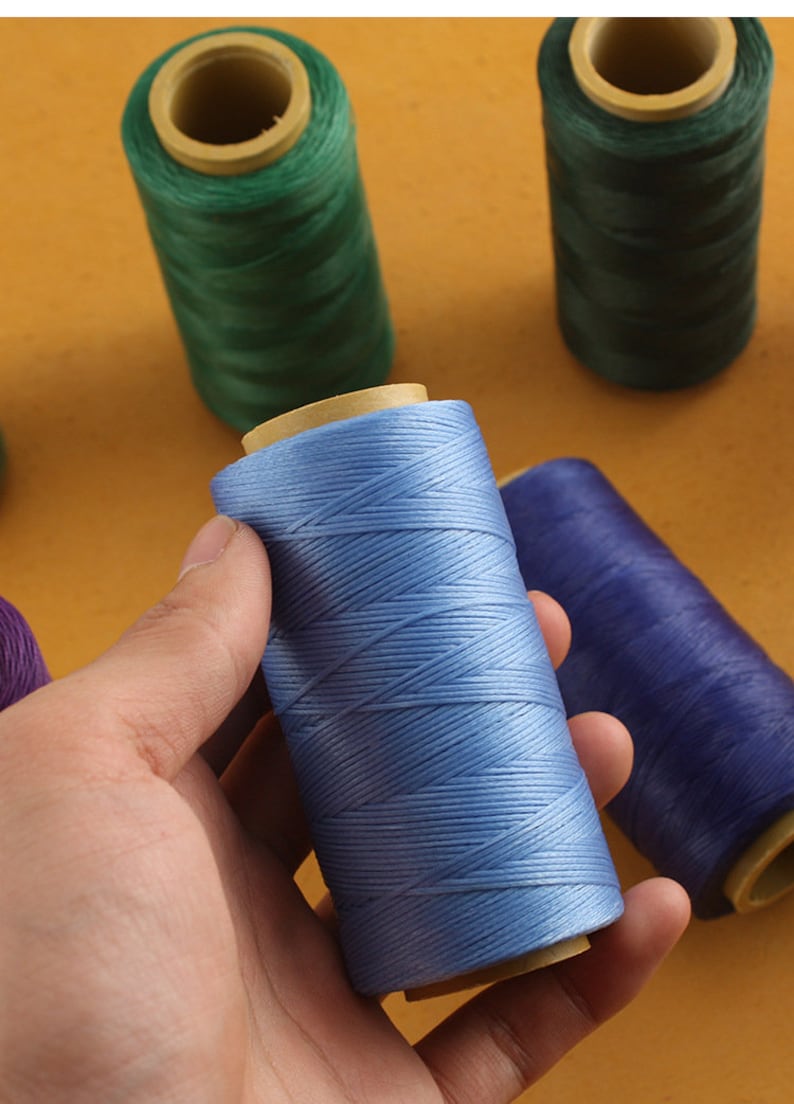 May include: A hand holds a spool of light blue thread, with other spools of green and purple thread in the background. The thread is tightly wound around a cardboard core. The background is a solid, warm yellow color.