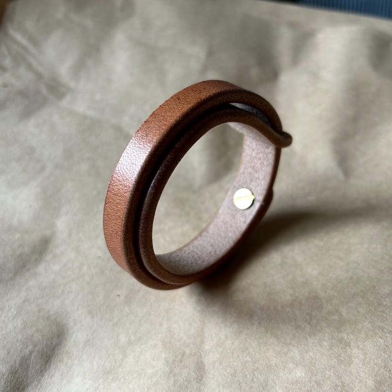 May include: A brown leather bracelet with a double-wrap design. The bracelet features a light brown interior and a small, gold-colored metal clasp. The leather has a smooth texture and a warm, natural color.