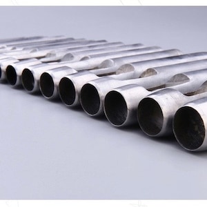 May include: A collection of silver-colored metal hole punches, arranged in a diagonal row. Each punch has a cylindrical body with a circular opening on one end and a curved, flattened section on the other. The tools are set against a light gray background.