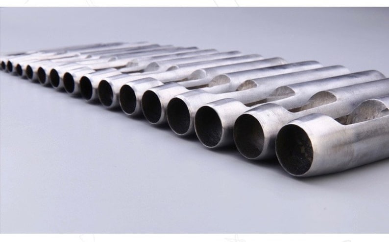 May include: A collection of silver-colored metal hole punches, arranged in a diagonal row. Each punch has a cylindrical body with a circular opening on one end and a curved, flattened section on the other. The tools are set against a light gray background.