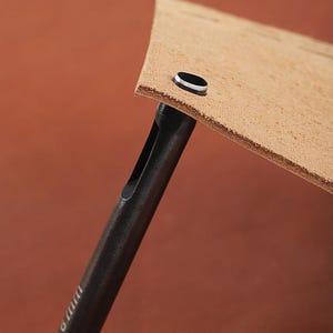 May include: Close-up of a leather crafting tool punching a hole in a piece of tan leather. The tool is black with a cylindrical shape and a hollow center. The leather is a light brown color.