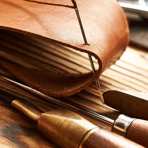 May include: Close-up of leather crafting tools, including a needle piercing through a piece of brown leather. Other tools with wooden handles and metal tips are visible, suggesting a leatherwork project.