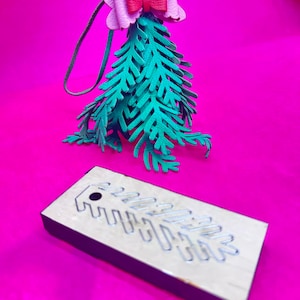 Könnte beinhalten: Handgefertigter Weihnachtsbaumschmuck mit einer grünen Lederbaumform, einer rosa Schleife und einer Lederschlaufe zum Aufhängen. Eine Holzstanze mit einem Blattausschnitt befindet sich im Vordergrund. Der Hintergrund ist leuchtend pink.