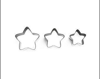 May include: Three star-shaped cookie cutters in varying sizes. The cutters are made of shiny, silver-colored metal and are arranged from largest to smallest. The star shapes have five points and are set against a plain white background.